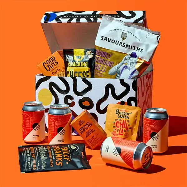A Fathers Day snack hamper displayed with a patterned gift box filled with crisps and treats, surrounded by canned drinks, biscuits and snack packs on a bright orange background.